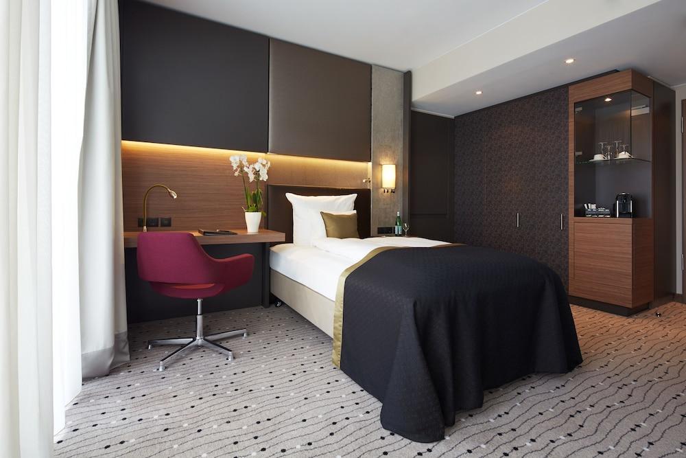 فندق ستيجنبرجر آم كانزليرمت برلين المظهر الخارجي الصورة The photo shows a modern hotel room featuring a single bed with a dark bedspread. The room has a contemporary design with a neutral color palette, accented by warm lighting. There's a small desk with a red chair positioned next to the bed. A cabinet 